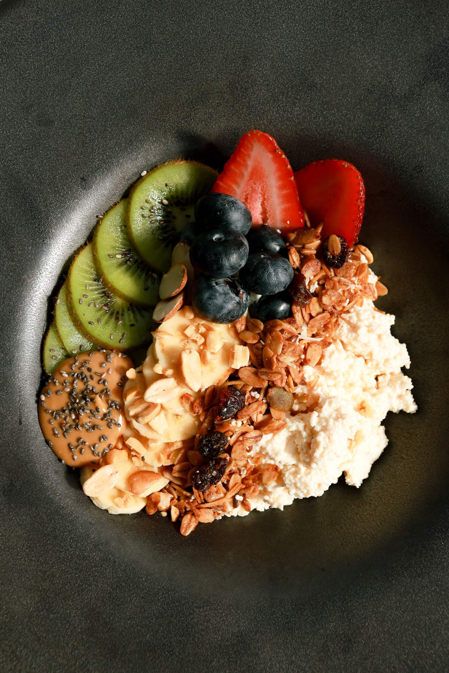 Bowl de queso cottage con kiwi, fresas, arándanos y granola sobre plato oscuro